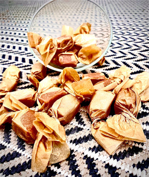 A rectangular box filled with 8 wrapped caramels. The label on top of the box says "caramels with sea salt," as well as "Scenic Made" and "small batch, big flavor." 3 caramels are sitting outside of the box. 2 are wrapped in natural color parchment paper,